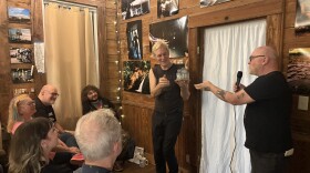 Scott Satterwhite and Aaron Elliot (aka Aaron Cometbus) speak to a seated audience inside the 309 Punk Project house in Pensacola, as Cometbus holds a small model of the home created by an artist in residence.