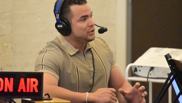 A man sits at a table with an "on-air" sign in front of him. The man is wearing a headphone and microphone set on his head. 
