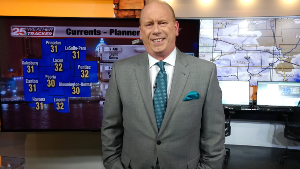 A man in a grey and blue suit stands behind a desk, with weather forecasting screens behind him.