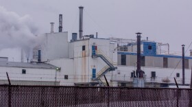 Industrial facility with white and blue exterior, multiple smokestacks emitting smoke, yellow staircases, and barbed wire fencing in the foreground on a cloudy day.