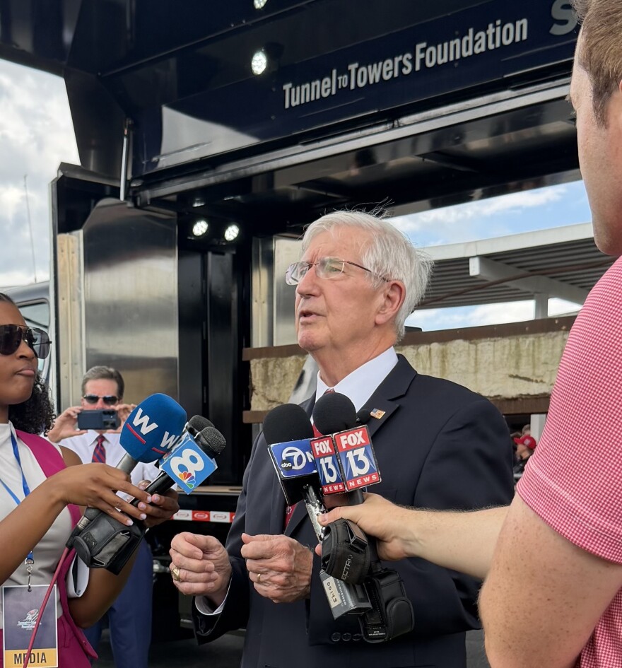 A white haired man in a blue suit clenches his fists as he is interviewed by reporters, with five mics in front of him