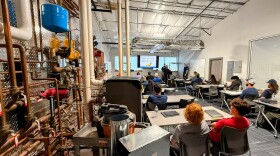 An image of a classroom taken from the back of the room. Several students sit at desks facing a projection screen. On the left side of the image the wall is covered with exposed piping and mechanical equipment
