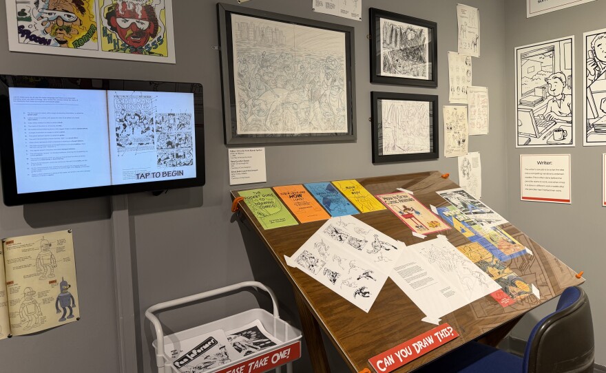 A desk with comic book pages. A sign below reads, "Can you draw this?"