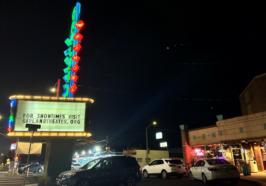 Cars sit parked near the front entrance of the Garland Theater, with the theater's sign lit up in colorful lights. 