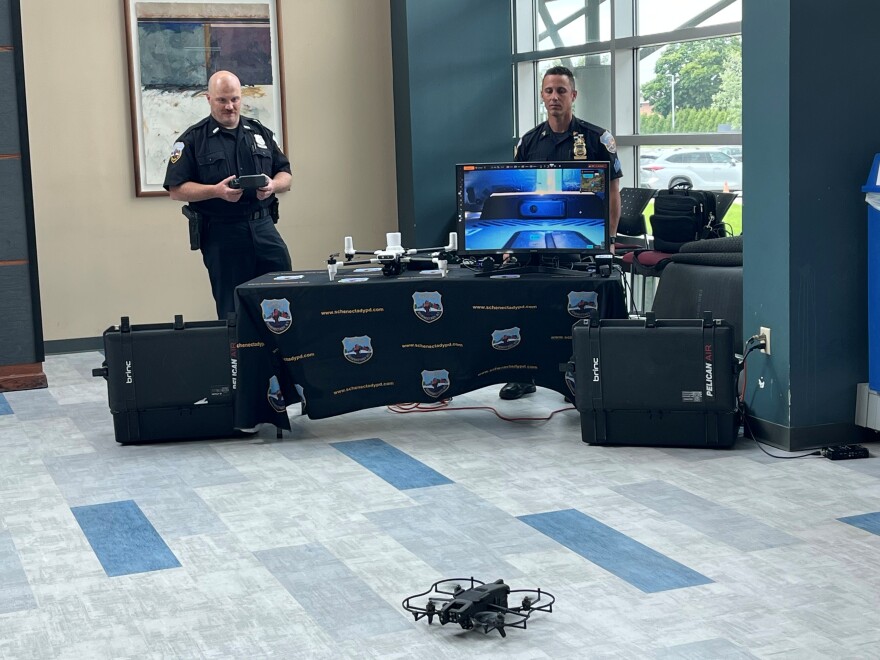Schenectady Police behind a table and a computer screen show off one of their drones during a demonstration at Hudson Valley Community College. The drone is on the floor in front of the table. 