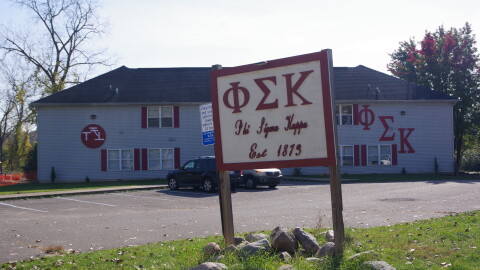 A large grey building sits in the background, with a sign in the foreground marking it as the Phi Sigma Kappa fraternity, with matching Greek letters. These letters can also be seen on the side of the building. 