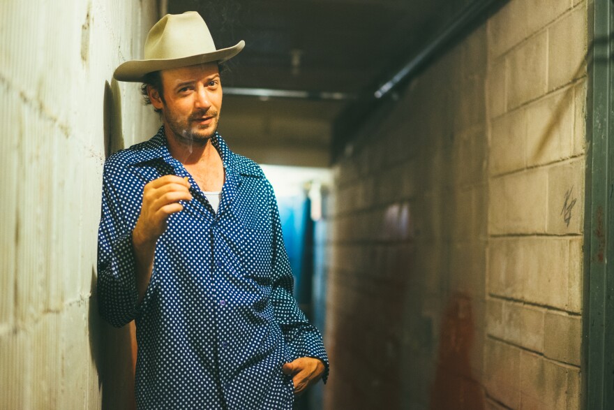 A man in a blue shirt and a white cowboy hat stands in a narrow hallway