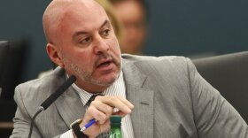A bald man with a beard at a microphone, wearing a tailored suit and a striped shirt