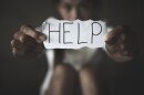 A woman sits alone holding out a piece of paper with the word HELP on it