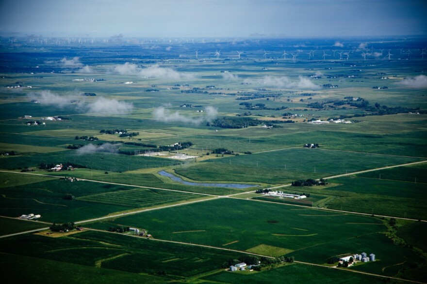 An aerial view of green farm fields and wind turbines on the horizon.