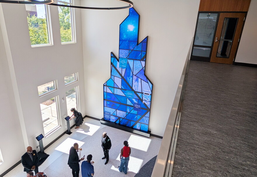 A balcony view looking down on the center's lobby, where several people are walking and talking. The focal point is an installation on the opposite wall. The two-story screen is shaped like jagged glass, and it projects a background that looks like blue stained glass, with four boxes containing written commitments in the foreground.