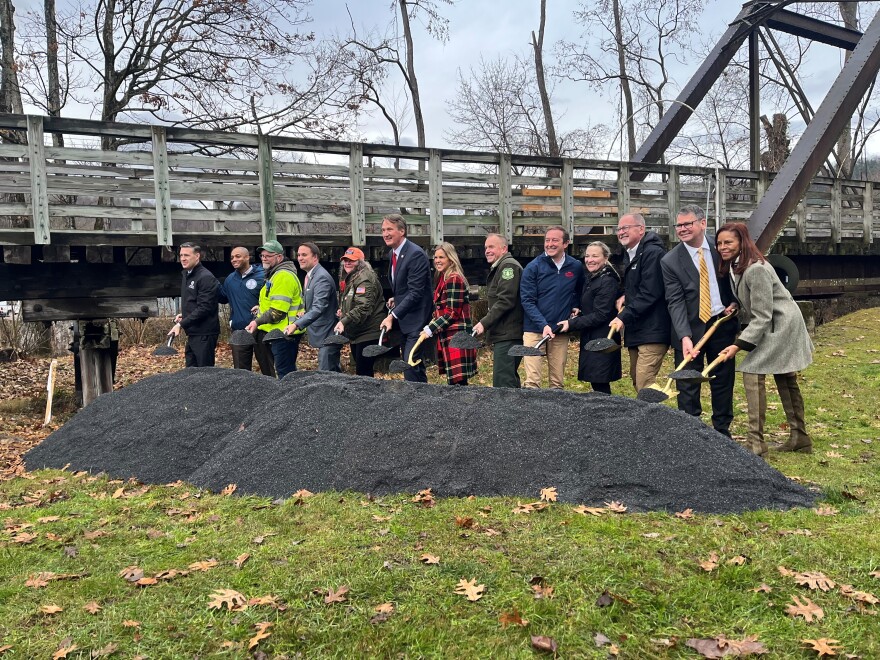 Gov. Glenn Youngkin (center), with 12 other federal, state and local officials, gather around a mound of grey crushed gravel, the same material used in the Virginia Creeper Trail. They hold shiny shovels, aimed at the mound. Behind them is a wooden bridge from the trail. 