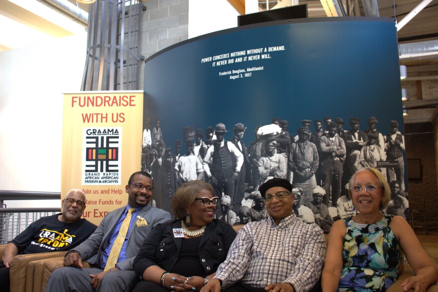 Five board members of the Grand Rapids African American Museum & Archives sit in chairs, smiling as they announce the museum has secured a new location. Behind them, a large black and white photo serves as the backdrop. To the left, a banner reads "FUNDRAISE WITH US" above the logo of the museum.