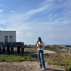 A woman stands holding a microphone with seated and standing audience members looking at her on the edge of the coast. 