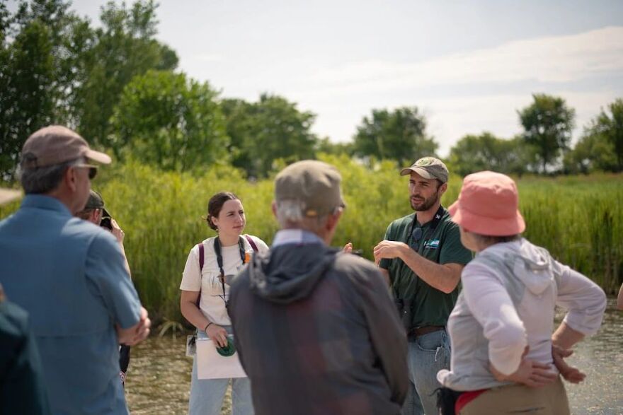 A small group of people in an open marshy area listen to one person talking