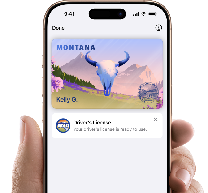 A hand holds an iPhone displaying a Montana Mobile ID in Apple Wallet. The digital ID shows a stylized landscape with mountains, flowers, and a blue bison skull, along with the name "Kelly G." A message below reads, "Driver’s License – Your driver’s license is ready to use."