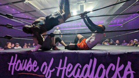 Three wrestlers compete around the ropes of a wrestling ring, with one falling over the edge. A crowd looks on in the background.
