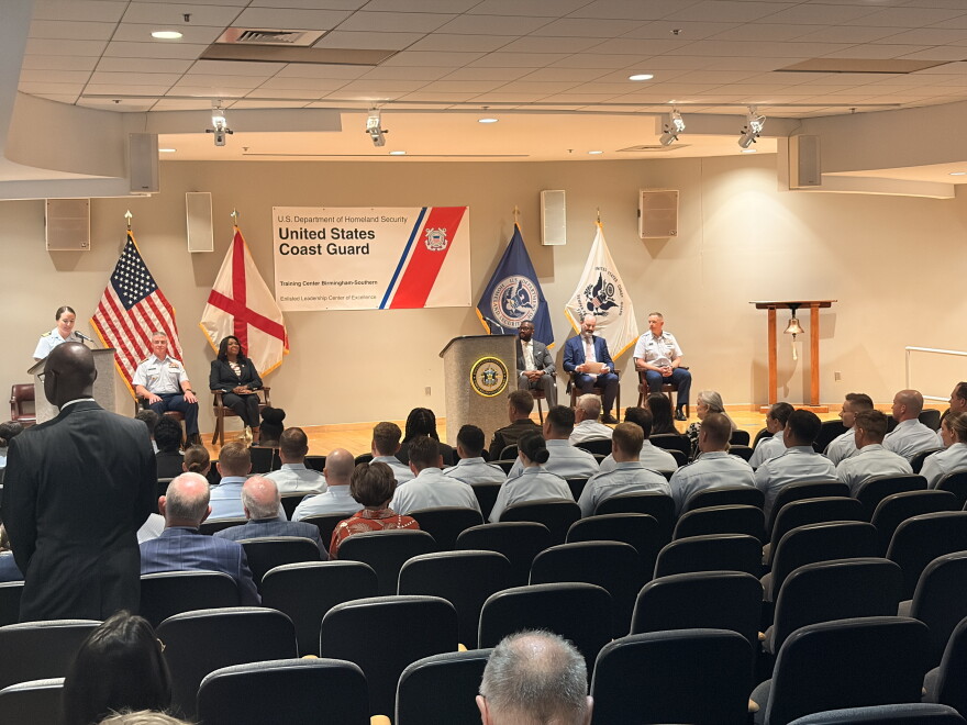 Coast guard graduates sit in the front two rows as an office speaks from a podium on the stage. 