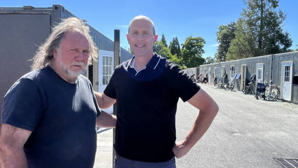 Two middle-aged men are standing next to each other outdoors. Behind them are shipping container homes. 