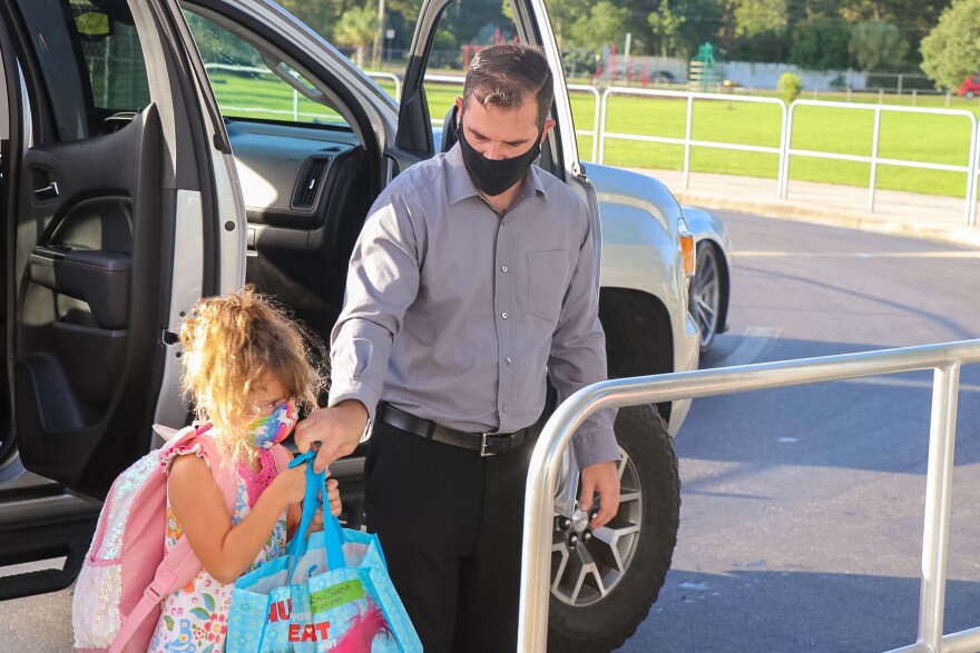 adult in gray button up shirt and mask escorts young girl in backpack and mask to front of a school.