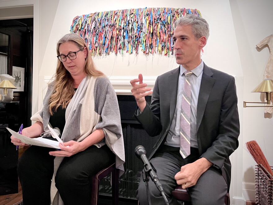A woman with long blond hair and thick black glasses holds a stack of papers and a pen as she listens to remarks made by a man beside her who has gray hair and wears a gray suit. They are seated on stools in front of a fireplace that is adorned with a colorful artwork made of bits of fabric.