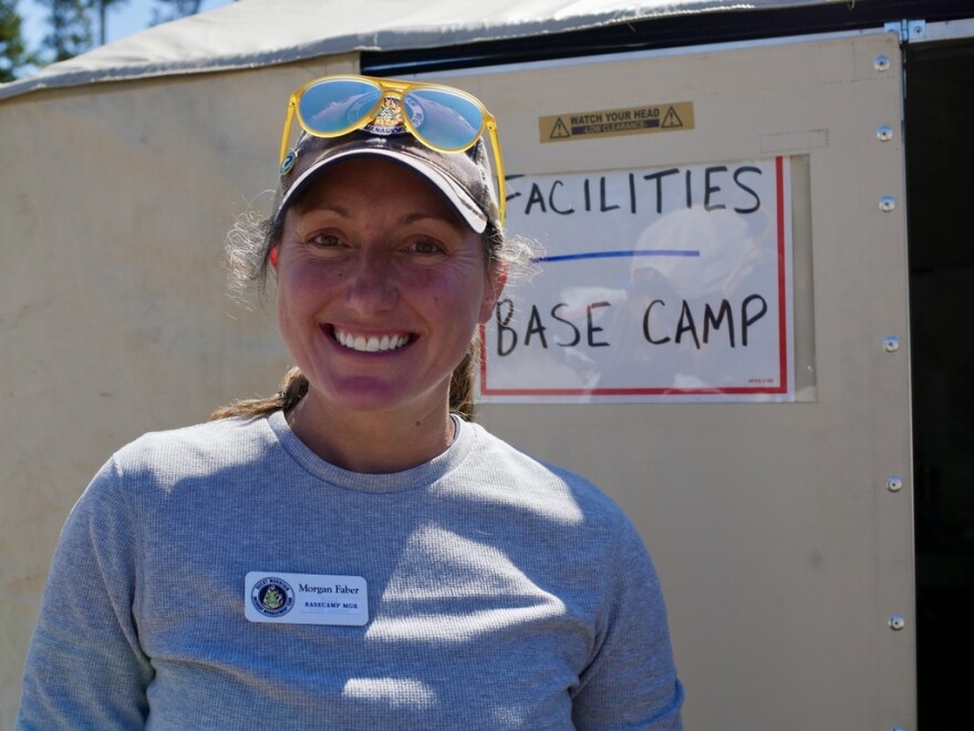 A woman hearing a brown baseball hat and a gray shirt stands in front of a yurt with the sign that says “Facilities / Base Camp.”