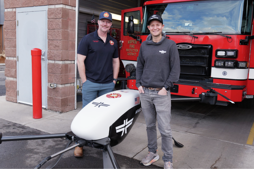 Two men stand in front of a fire engine and beside a small drone, about the size of a bicycle