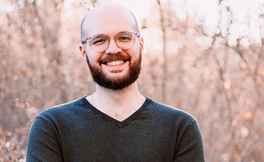 A bald man with a brown beard is wearing glasses and a dark sweater and standing outside with his arms crossed against his chest.