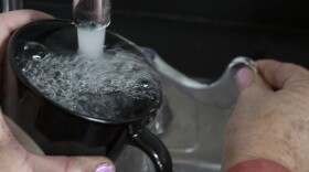 A woman's hand holds a black coffee cup beneath a faucet. Her other hand is turning the lever to run the water.