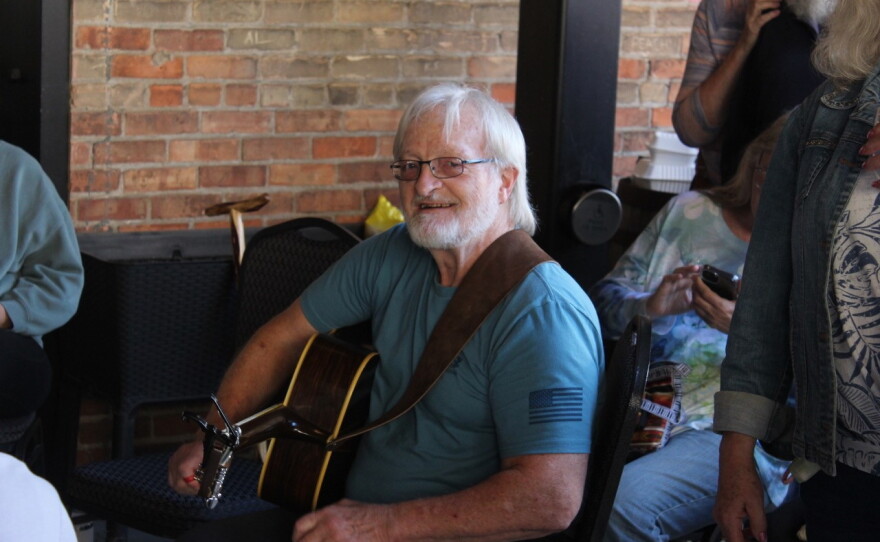 A man sitting with a guitar looks and smiles at the camera.
