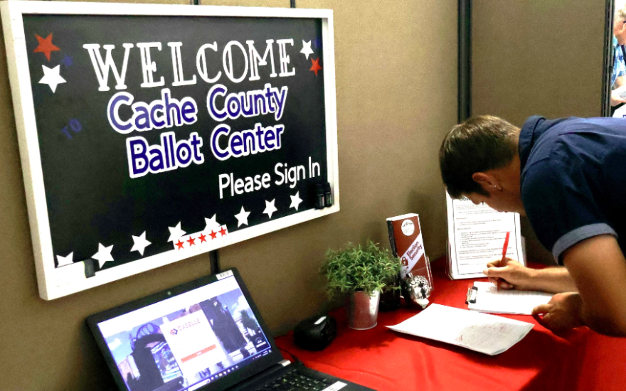 Visitor Chris Barnes signs in to the ballot center. Anyone entering the building must record their name, as well as the date and time of their visit. The sign above him reads "Welcome. Cache County Ballot Center. Please sign in."