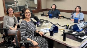 Four smiling people wearing headphones sit at a table in a radio talk studio: A woman front left has shoulder-length dirty blonde hair and is wearing glasses and a grey top; a woman front right has short dark curly hair and is wearing glasses and a black and white striped dress; a man back left has short brown hair, a brown beard and mustache, and is wearing glasses and a blue polo shirt; a woman front right has long brown hair and is wearing a blue floral blouse and light blue sweater