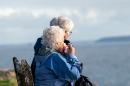 An older couple sits on a wooden bench, looking out across a body of water