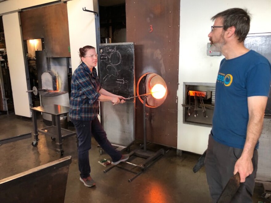 Lena Quade works glass in the reheating chamber while talking with fellow Glass Center instructor Robert Warden.