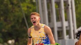 A boy in a track uniform crosses a finish line with a ribbon breaking over his torso.