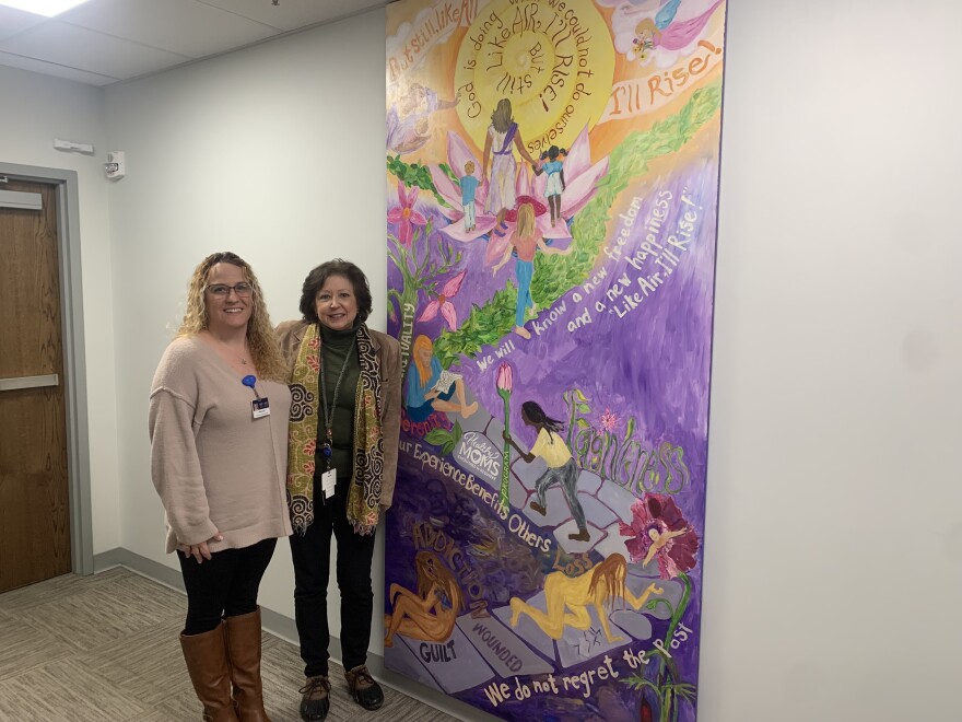 From left to right: Michelle Coyle, Healthy MOMS Case Manager and Doula; and Maria Kolcharno, Healthy MOMS Supervisor stand next to a mural painted by moms in the program.