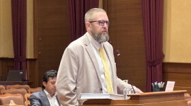 A bearded man in a blazer and tie speaks at a podium in a courtroom, with another man sitting behind him.