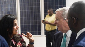 Woman in flower-patterned blouse speaks with two men in suits in front of solar panels