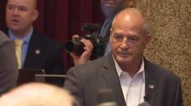 Gary Pinkel is seen in front of microphones. There is a photographer in the background with a man in a suit. Pinkel has a neutral expression.  