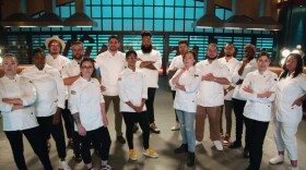 A group of 15 chefs in white jackets pose while looking at the camera in a kitchen setting.