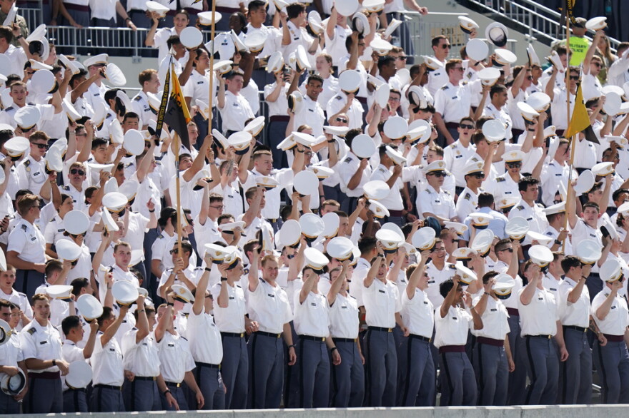 Group of Army cadets in white uniform tops and white caps salute by raising their caps to a the crowd at a stadium 