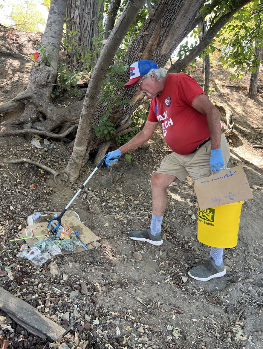 An older man wearing a red t-shirt, glasses, and a baseball cap picks up trash in the dirt using a grabber. 