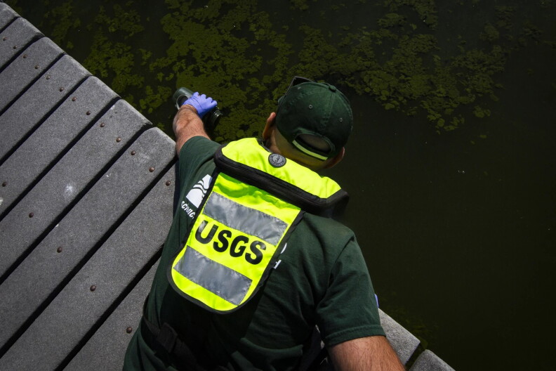 A man on a dock scoops up greenish water into a water bottle.