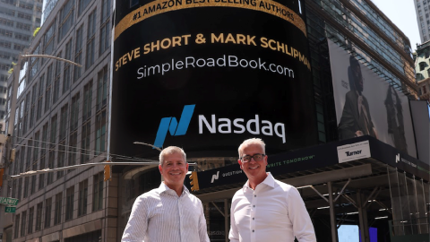 Two men in button up shirts pose for a photo in front of an advertisement display for Amazon best selling authors Steve Short and Mark Schlipman 
