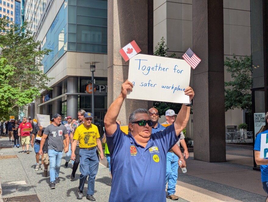 People, mostly men, walk on a downtown sidewalk. A man in the front carries a handmade sign that reads, "Together for a safe workplace," with American and Canadian flags taped onto each corner.