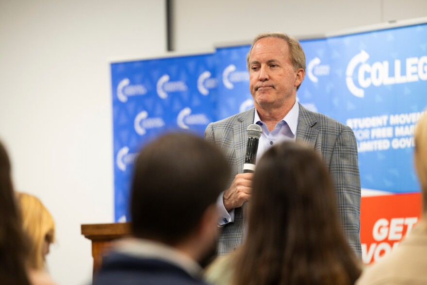 A gray-haired man holds a microphone at an event where people are gathered, listening