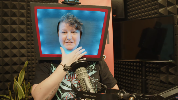 A white person with short, dirty blonde curly hair sits in a studio at a mic. They are wearing a ‘Hazbin Hotel’ t-shirt featuring Vox, a character with a TV for a head. They are also wearing a foam cosplay piece - the same TV head - that they made for a cosplay. 
