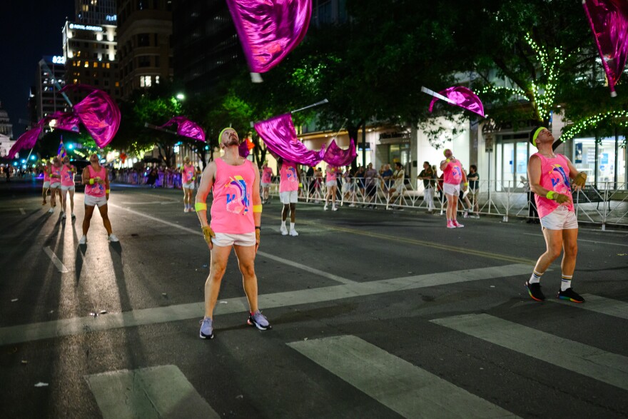 Flag twirlers wearing white shorts and neon pink tan tops with neon headbands and wrist bands march down Congress Avenue during the Austin Pride Parade. Here the people are looking up at their metallic pink flags that they threw up in the air.