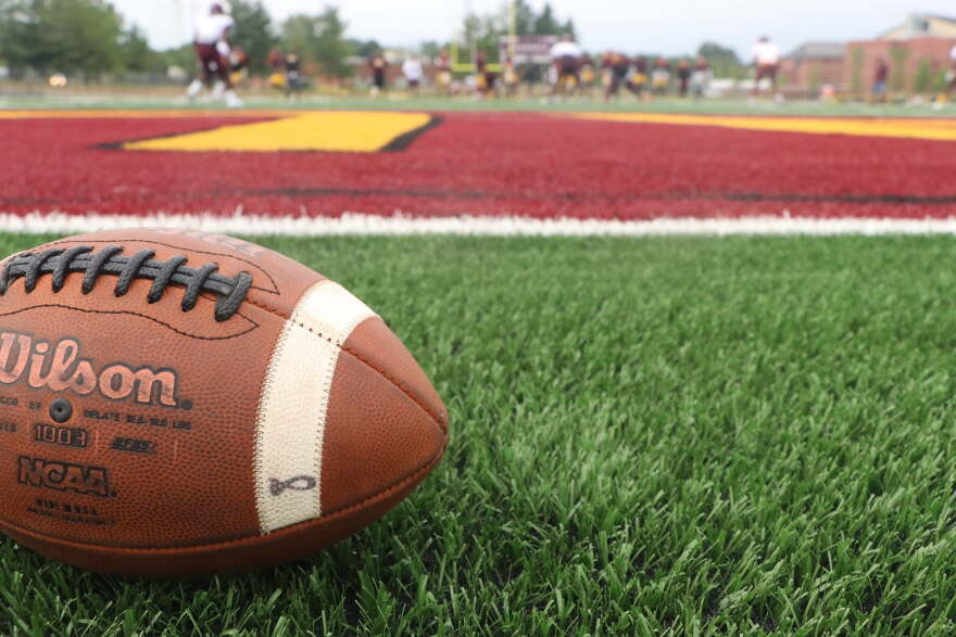 A football sits in the foreground of a green field, with red and gold markings on the field.