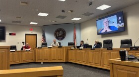 Four people sit at a daias in the Jefferson City Municipal Building meeting room. Three men are to the left, and one woman is to the right. A council member's face appears on a screen behind the woman; he and few others are joining the meeting on Zoom.